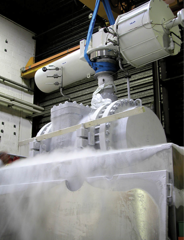 Industrial valve in a factory sprayed with a frost-like substance, likely indicating operation in a cryogenic process. The valve, connected to a pipeline, is mounted on a metal structure. Background includes large cylindrical tanks and metal shutters.