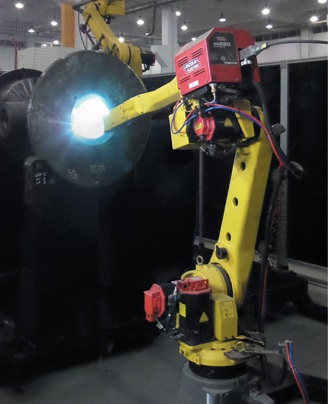 A yellow industrial robotic arm equipped with a red welding machine is operating in a factory. The robotic arm is positioned near a large cylindrical component, emitting a bright blue light indicative of welding or cutting. The background shows a high-ceiling industrial space.
