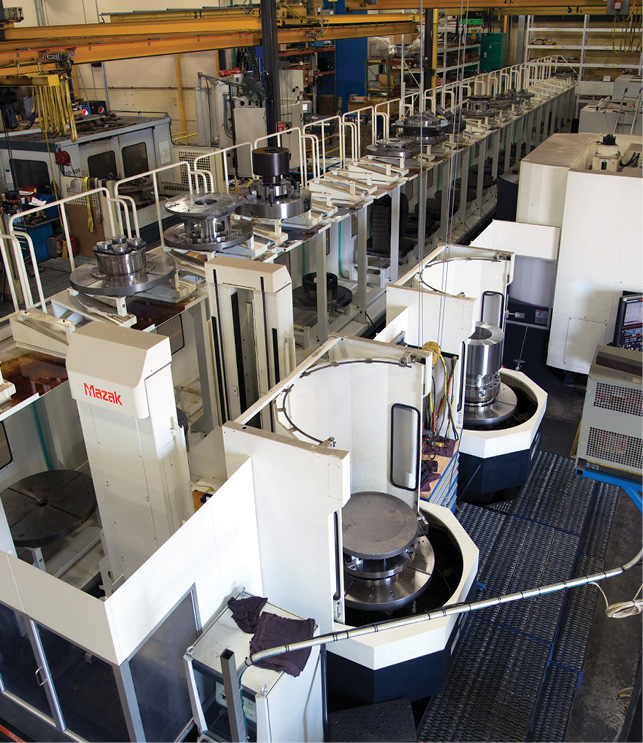 Bird's-eye view of an industrial manufacturing facility showcasing a row of large CNC machines. The setting features various machinery components, control panels, and safety railings. A yellow crane can be seen overhead, and some textiles are draped on the equipment.