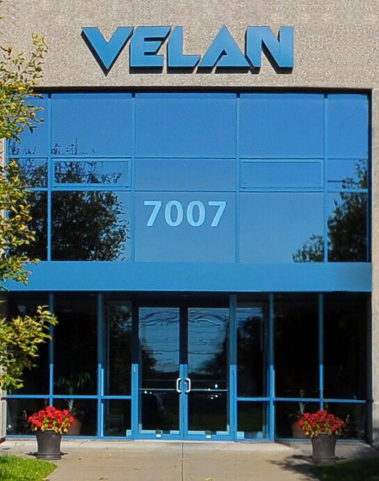 A modern building entrance with large blue-tinted windows and the number "7007" displayed prominently above the doors. The word "VELAN" is written at the top. There are two potted plants with red flowers on either side of the entrance walkway.