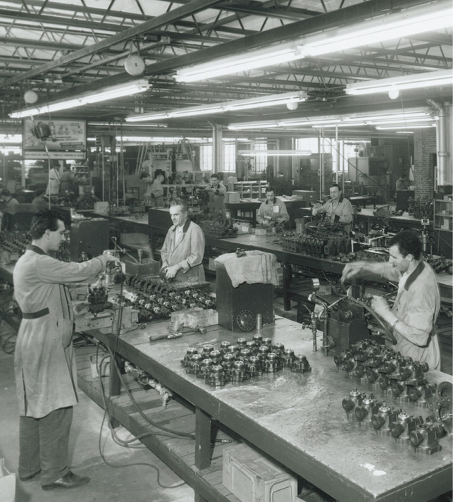 Black and white image of a factory workshop. Multiple workers, mostly men, are busy at large tables filled with mechanical parts. The space is lit with overhead fluorescent lights. Shelves and additional parts are visible in the background.