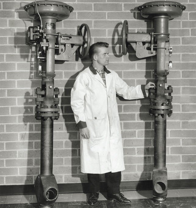 A person wearing a lab coat and dark pants stands between two large industrial valves attached to a brick wall. They appear to be inspecting or operating the valve on the right, with one hand on the valve wheel and the other resting on part of the mechanism.
