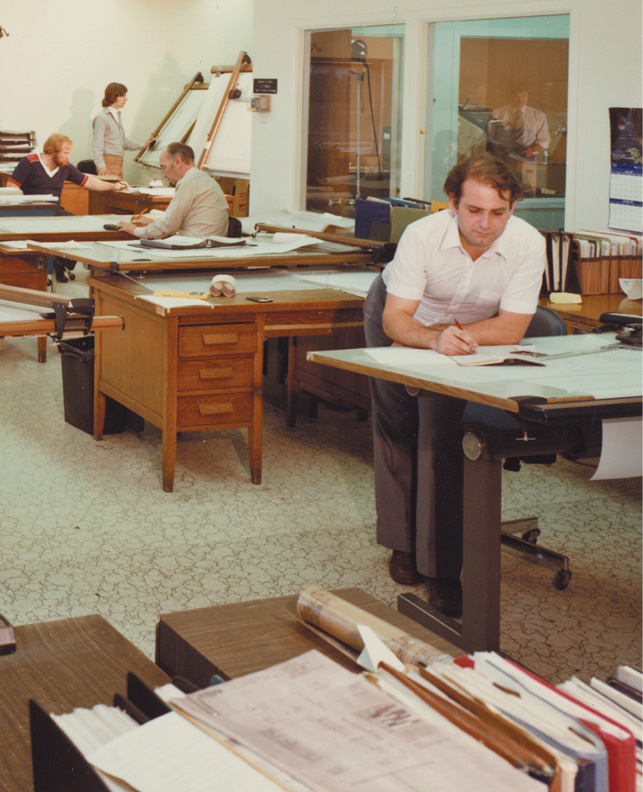A group of people work in an office space with large wooden desks covered in papers. One man stands and writes on a drafting table, while another sits nearby. A woman and another man are seen in the background working at their desks. The room has a professional atmosphere.