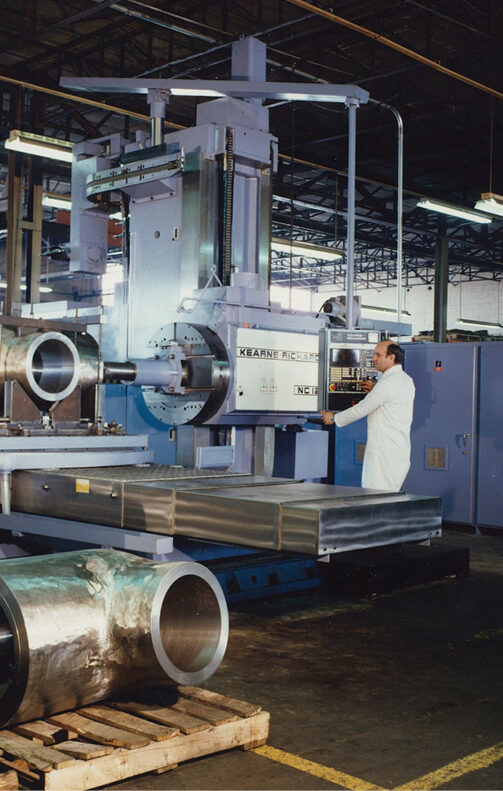 A man in a white lab coat operates a large industrial milling machine in a factory. Nearby, several large cylindrical metal parts are placed on the floor. The factory has high ceilings and visible structural beams. The machine is labeled with "KEARNEY & TRECKER.