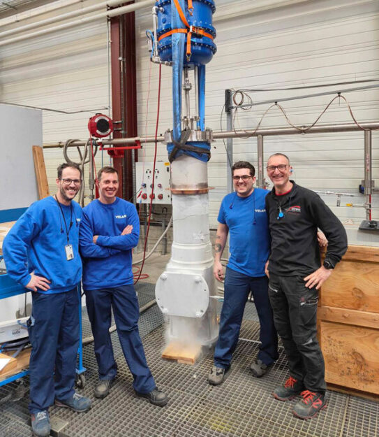 Four people are standing in a workshop next to a tall industrial valve. Two of them are wearing blue uniforms, and the other two are in dark clothing. They are all smiling at the camera, posing with the valve that has blue and silver components.