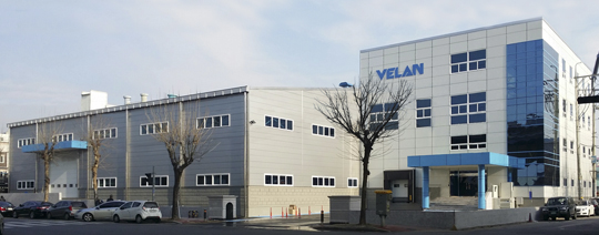 A modern industrial building with large glass windows and a sign reading "VELAN." Several parked cars are visible in front of the structure, with a clear sky overhead. The architecture is clean and geometric.