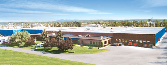Aerial view of a large industrial building with a blue and brown exterior, set amidst a spacious green lawn. Trees surround the property, and a clear blue sky is visible in the background.
