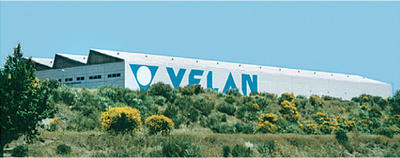 A large industrial building with a white facade displaying the word "VELAN" in blue letters. The structure is partially obscured by greenery and yellow flowering bushes, under a clear blue sky.