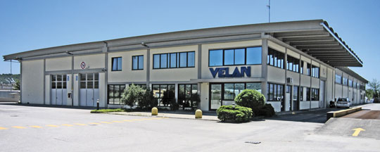 A large industrial building with multiple windows and two main entrances. The facade is white with a blue sign reading "VELAN." The area in front is paved, with some greenery near the entrance. The sky is clear and blue.