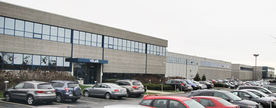 A large industrial building with a long row of windows on the upper floors. Numerous vehicles are parked in the adjacent parking lot. The sky is overcast.