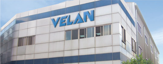 The image shows a building with a large sign that reads "VELAN" in blue letters. The structure features a modern design with reflective windows and light-colored exterior panels.