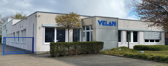 A single-story office building with a white exterior and blue accents. The word "VELAN" is prominently displayed on a sign. The building is surrounded by hedges, a tree, and a blue fence on the left. The sky is partly cloudy.