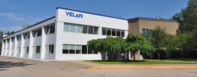 A large white and beige building with the word "VELAN" on the front. It features multiple windows and a small entrance surrounded by greenery. The sky is clear and the area appears quiet and well-maintained.
